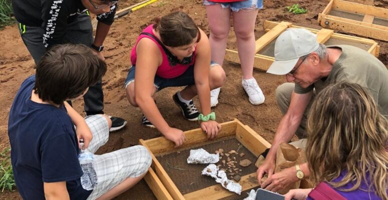 Youth participate in artifact sorting. They lean over screens placed on the ground to look at artifacts.