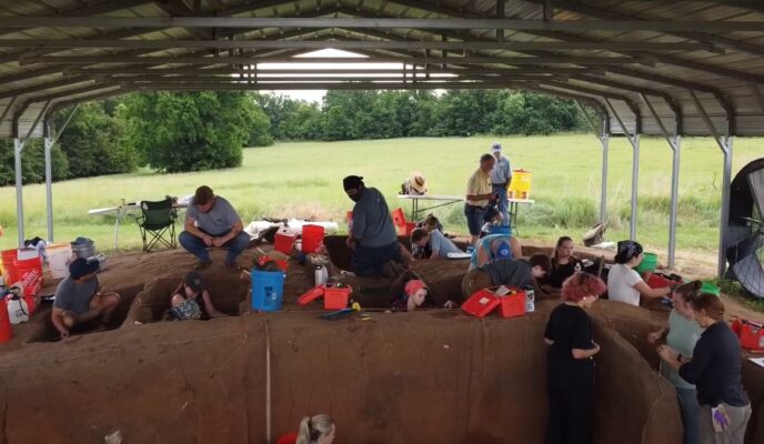 A group of students and excavation participants digging in units under a covered pavilion.